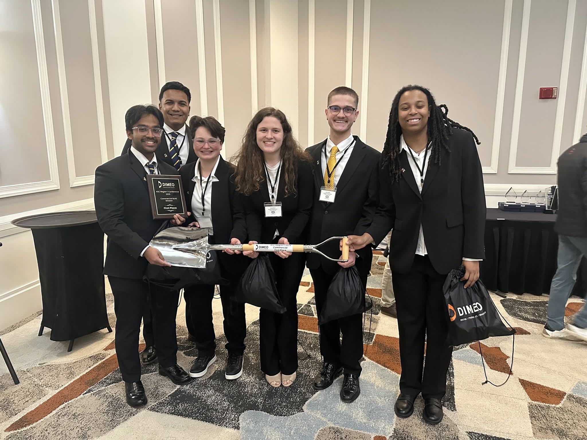 Students in suits holding giant shovel trophy